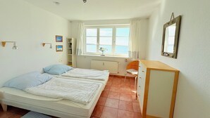 Ett sovrum med två sängar, ett skrivbord, en stol, ett element, ett fönster med gardiner och en spegel på väggen.