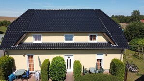 A two-story house with a dark tiled roof, a beige exterior, and a front entrance with a door labeled "HEUS". There are two windows on the upper floor and a small porch area with a table and chairs. - Apartment Seelenfrieden 3 - with balcony for 4 persons - Cozy vacation apartment with balcony (Alt Reddevitz)
