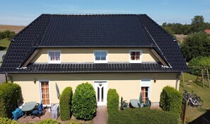 A two-story house with a dark tiled roof, a beige exterior, and a front entrance with a door labeled "HEUS". There are two windows on the upper floor and a small porch area with a table and chairs. - Apartment Seelenfrieden 3 - with balcony for 4 persons - Cozy vacation apartment with balcony (Alt Reddevitz)