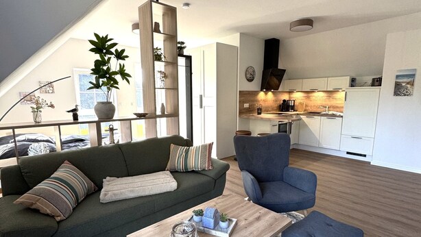 A modern living room with a sofa, armchair, and coffee table, a kitchen in the background, and a staircase leading to an upper level. - Vacation apartment Kajüte 2 - close to the harbor, with balcony - Fully equipped (Poseritz)