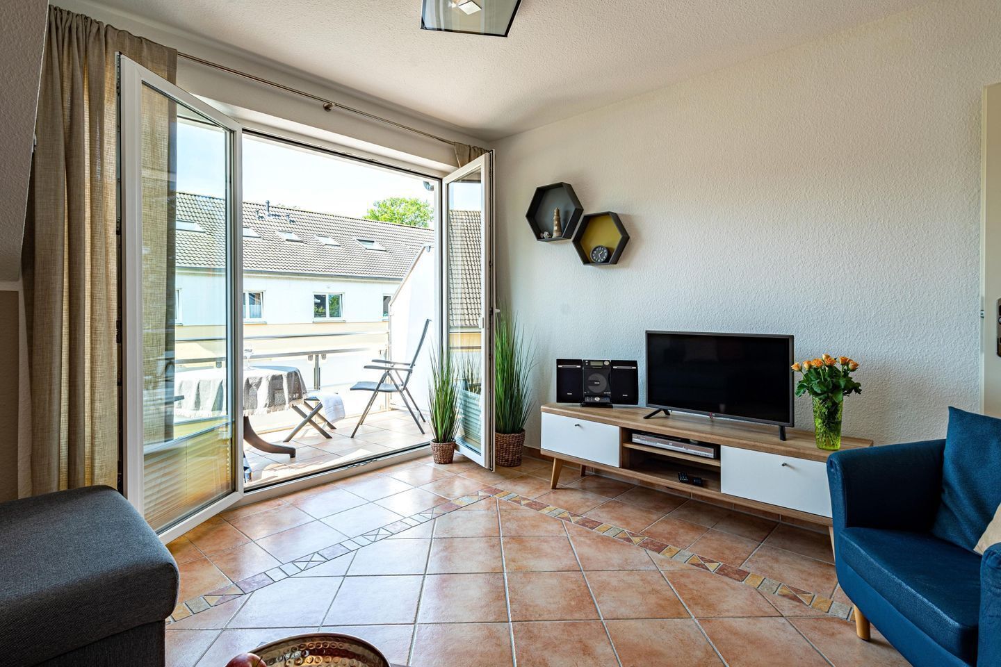 A living room with a TV, speakers, and a plant. There is a sliding glass door leading to a balcony with outdoor furniture.