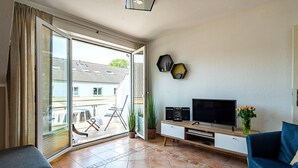 Ein Wohnzimmer mit Fernseher, Lautsprechern und einer Pflanze. Eine Glastür führt auf einen Balkon mit Gartenmöbeln.