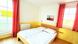 A single bed with a wooden frame and white bedding in a room with yellow walls, a window with red curtains, and a wall-mounted light fixture. - Fewo Seelenfrieden 6 - top for 2 persons and dog - vacation apartment in Alt (Alt Reddevitz)