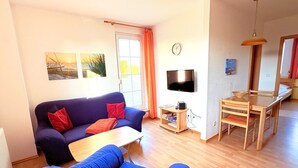 A living room with a sofa, a coffee table, a TV, and a dining area with a table and chairs. - Fewo Seelenfrieden 6 - top for 2 persons and dog - vacation apartment in Alt (Alt Reddevitz)