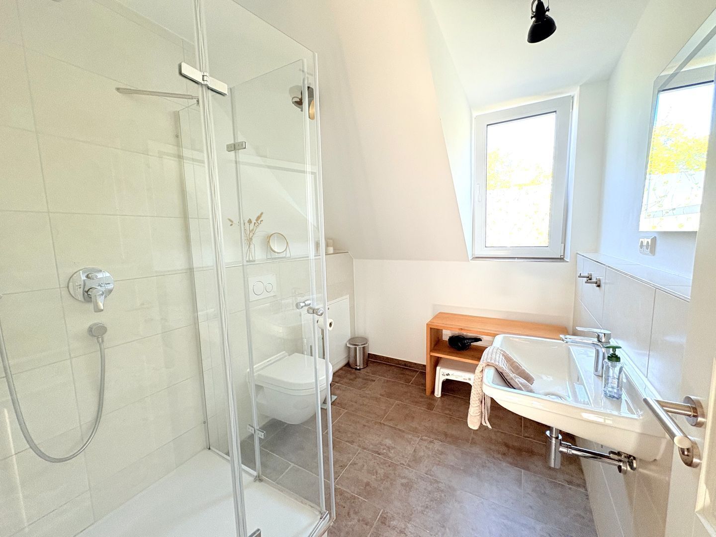 Ein modernes Badezimmer mit einer gläsernen Duschkabine, einer weißen Toilette, einer Holzbank und einem Waschbecken mit Spiegel.