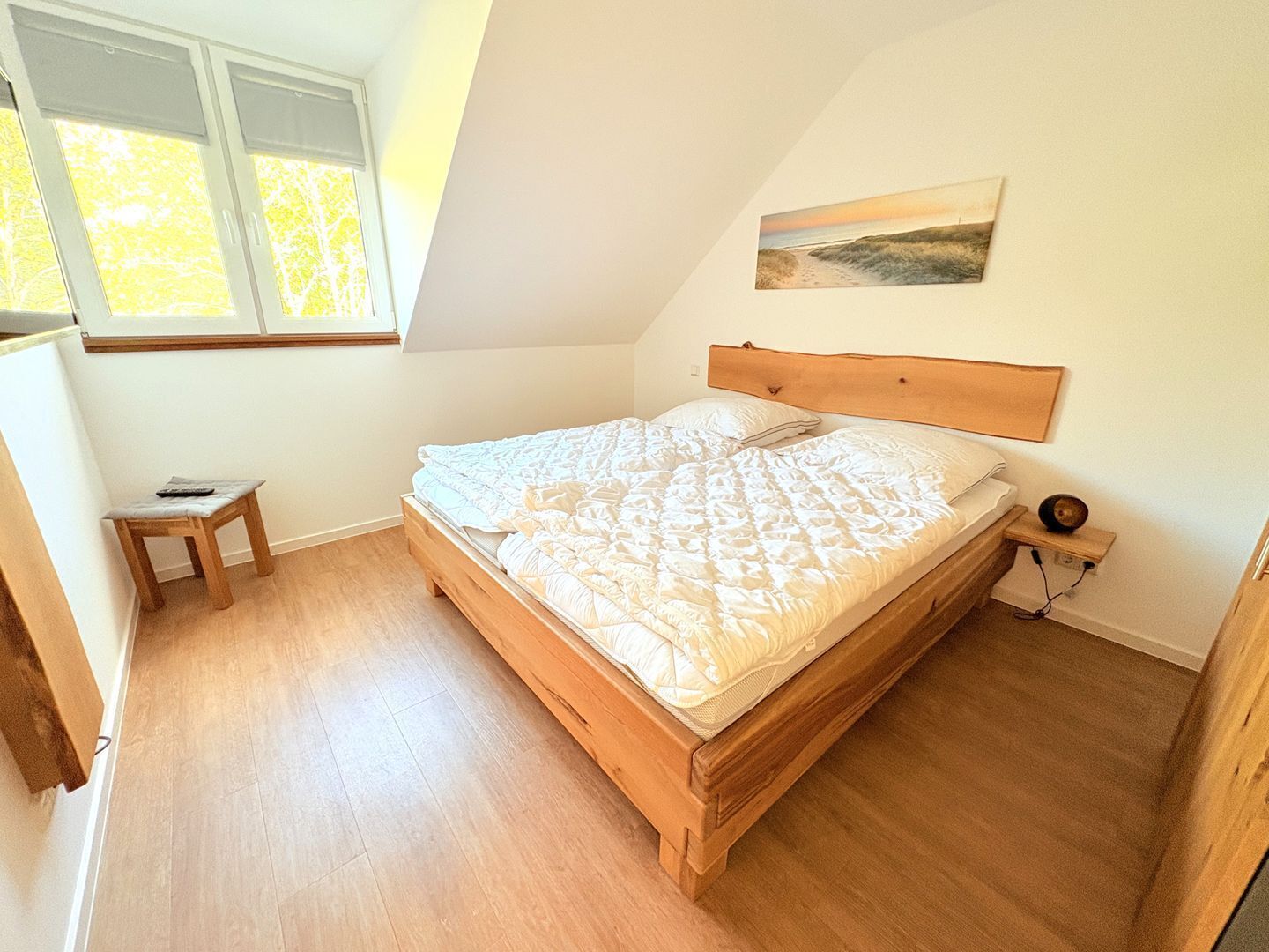 A bedroom with a wooden bed, a small bedside table, a nightstand, and a window with blinds.