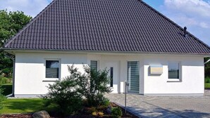 Uma casa moderna térrea com telhado escuro de azulejos, paredes externas brancas e um pequeno jardim frontal com um arbusto e um caminho de acesso pavimentado.
