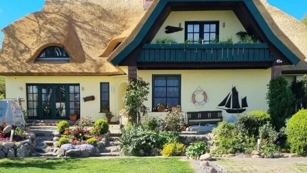 Una casa tradicional con techo de paja, un pequeño jardín con rocas y plantas, y un mural de un velero en la pared.