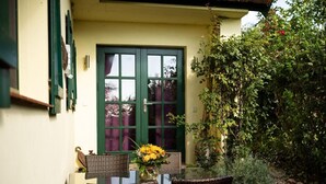 Un patio con sillas de mimbre y una mesa, una puerta verde y un jardín con diversas plantas.