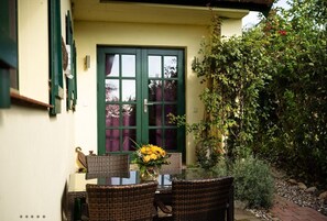 Une terrasse avec des chaises et une table en osier, une porte verte et un jardin où poussent différentes espèces de plantes.