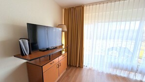 Hotellihuone, jossa on puinen työpöytä, taulu-tv, lamppu ja suuri ikkuna, jossa on läpikuultavat verhot.