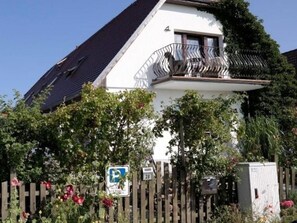 Ein zweigeschossiges Haus mit Balkon, umgeben von einem Holzzaun und blühenden Pflanzen.