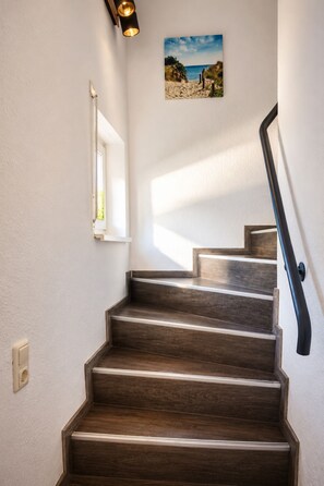 Un escalier avec des marches en bois et une rampe noire, menant à un palier où est accrochée une image représentant une scène de plage.