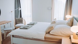 Ein ordentlich bezogenes Bett mit Kissen und gefalteten Handtüchern, ein Nachttisch mit Lampe, ein Sessel und ein kleiner Schreibtisch ebenfalls mit Lampe.
