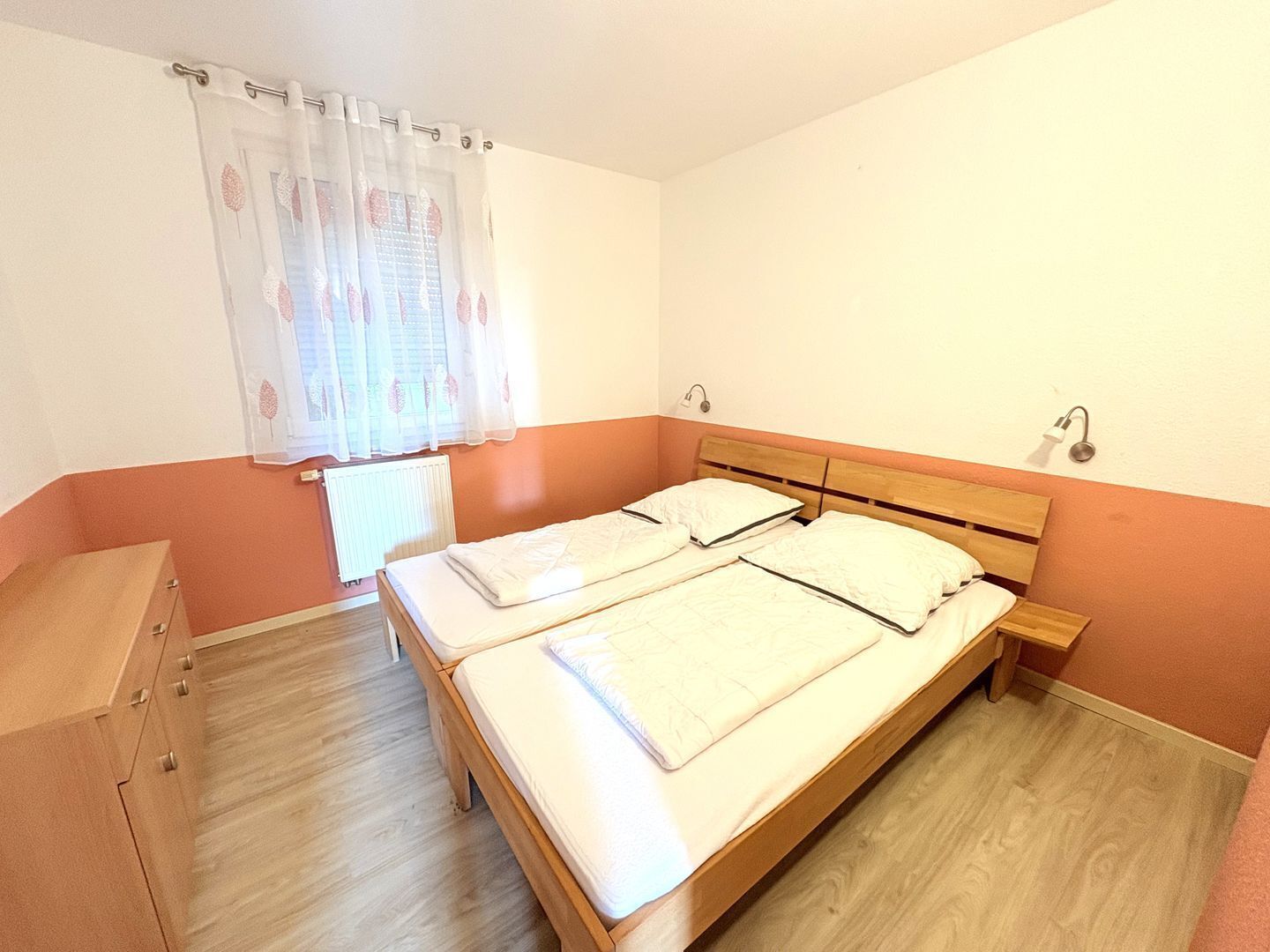 A double bed with a wooden headboard, a bedside table, a window with a sheer curtain, and a radiator.