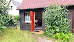 A wooden cottage with a red roof, a garden with a large bush, and a patio with chairs.