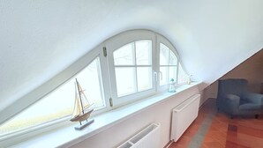Een kamer met een schuine kap, een raam met een model zeilboot, een radiator en een blauwe stoel.