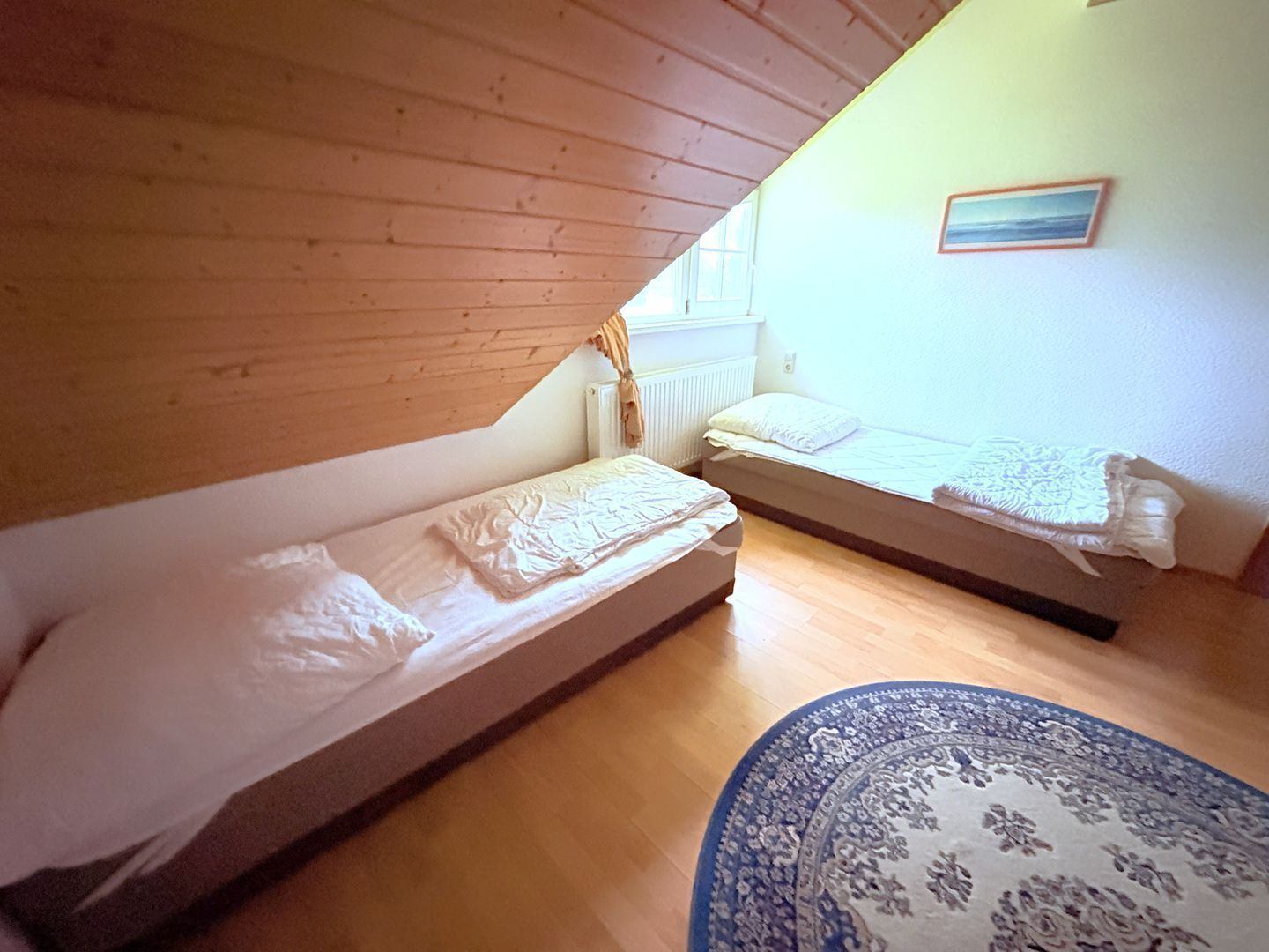 Um quarto pequeno e acolhedor com duas camas de solteiro, teto de madeira, uma janela e um tapete estampado.