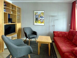 Ein Wohnzimmer mit Fernseher, zwei Sesseln, einer Couch, einem kleinen Tisch und einem Bücherregal.
