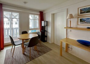 Ein Zimmer mit einem runden Tisch und Stühlen, einem Bücherregal, einer Lampe und Blick durch große Fenster.