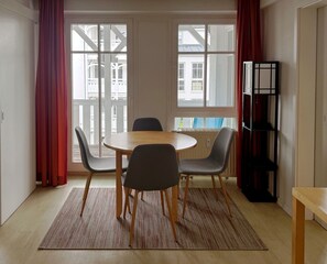 Una habitación con una mesa redonda de madera, cuatro sillas, una estantería y ventanas grandes con cortinas rojas.