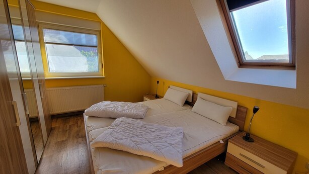 Een slaapkamer met een schuine plafond, een bed met witte beddengoed, een nachtkastje en een raam.