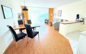 Una habitación de hotel moderna con zona de comedor, sofá, estación de café y un amplio ventanal con cortinas.