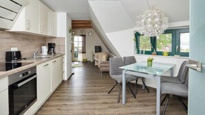 A modern kitchen with white cabinets, a dining area with a glass table and grey chairs, and a large window with a view of greenery. - Apartment 3 in the Zeesboothaus- balcony, view, sauna - Apartment 3 in the Zeesboothaus (Middelhagen)