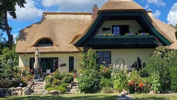A traditional thatched-roof house with a sloped roof, a small balcony with flowers, and a well-maintained garden with steps leading to the entrance. - Apartment 3 in the Zeesboothaus- balcony, view, sauna - Apartment 3 in the Zeesboothaus (Middelhagen)