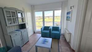 Quarto com cozinha compacta, duas poltronas azuis, uma mesinha e uma janela grande com vista para casas.