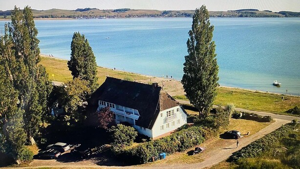 Uma casa com telhado de palha, rodeada por árvores, perto de um corpo d'água.