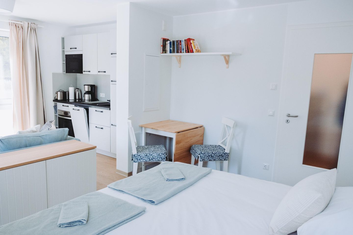 Ein modernes Hotelzimmer mit Bett, Schreibtisch, Stuhl, einer kleinen Küchenzeile und einem Bücherregal.