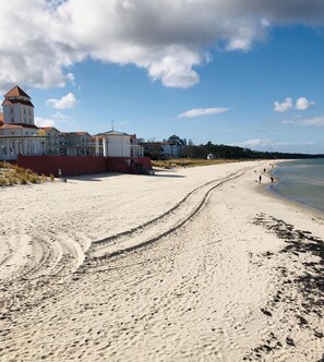 En strand med en sandstig som leder ner till vattnet, byggnader i bakgrunden och en klar himmel.