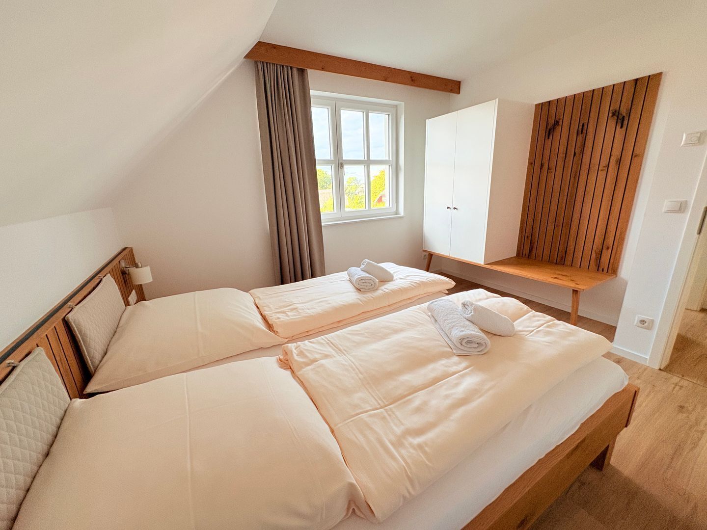 A room with two beds, a window with curtains, a wooden bench, and a wardrobe.