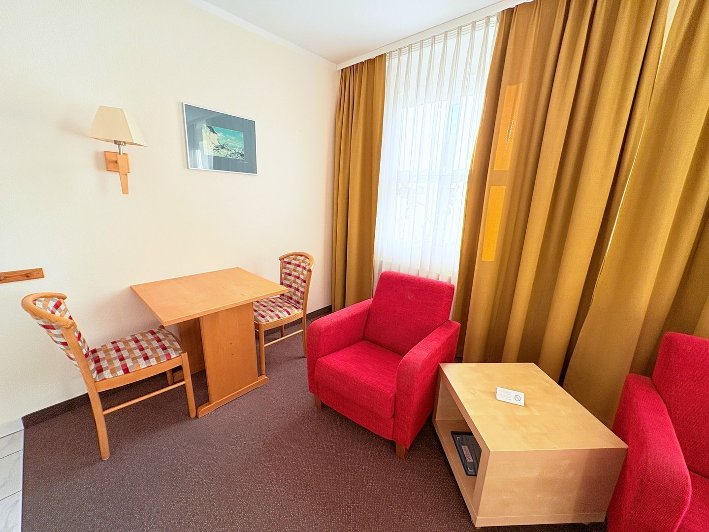 Habitación de hotel con una mesa de madera, dos sillas a cuadros, un sillón rojo, un pequeño armario de madera, una lámpara de pared y un cuadro enmarcado.