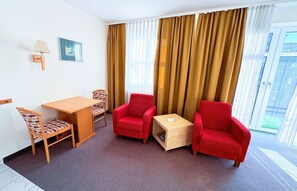 A hotel room with a wooden table, two red armchairs, a small wooden side table, a wall-mounted lamp, and a framed picture. - Apartment 8 in Haus Hühnergott - great for small families - Holiday apartment 8 - (Sellin)