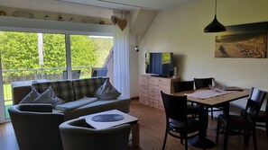 A room with a sofa, dining table, chairs, a TV, and a balcony with a view of greenery. - Boddenhof Apartment 52 - Balcony, dog-friendly - Apartment 52 - (Bergen)