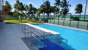 Piscina com grades de metal, cercada por uma cerca e palmeiras, com vista para a praia e o oceano.