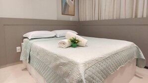 Uma cama bem arrumada com colcha acolchoada, uma pequena planta em vaso e uma toalha enrolada na cabeceira.