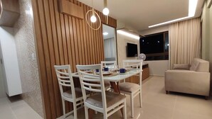 Uma sala de estar moderna com área de jantar, apresentando uma parede de destaque em madeira, uma televisão de tela plana e um sofá bege.