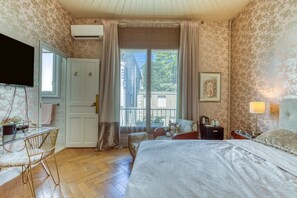 Ein Schlafzimmer mit Bett, Schreibtisch, Stuhl, einem Fenster mit Vorhängen, einem an der Wand montierten Fernseher und einem gerahmten Bild.