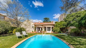 Una piscina con sillas de descanso, un porche cubierto y una casa con techo rojo al fondo.