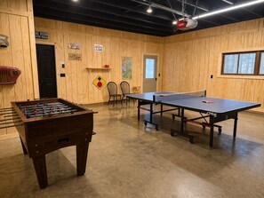 A room with a foosball table, ping pong table, chairs, and a door with signs on the wall.