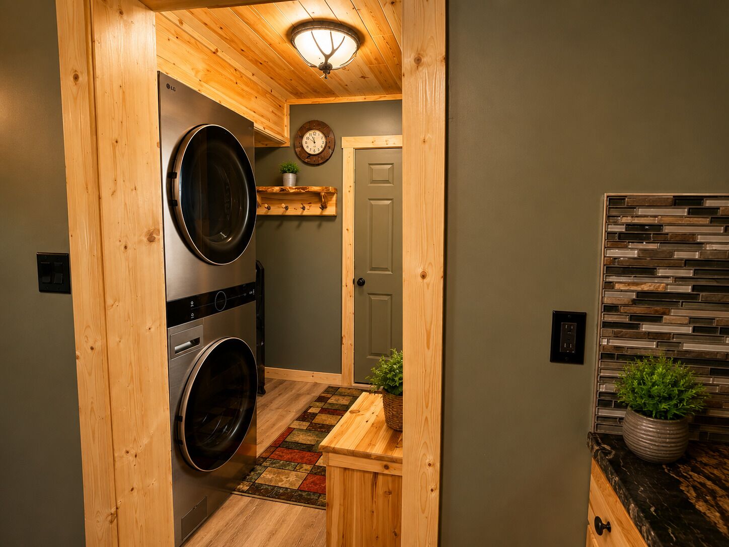 Une buanderie équipée de deux machines à laver et de deux sèche-linge à chargement frontal superposés, d’un plafond en bois avec un luminaire, d’une petite étagère ornée d’une plante et d’une porte donnant sur une autre pièce.
