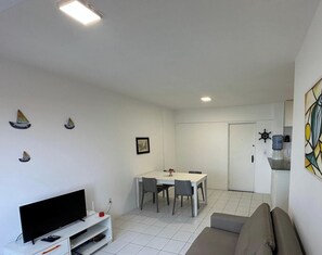 Een woonkamer met een televisie, een bank, een eettafel met stoelen en een keukenhoek met een waterdispenser.