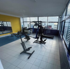 Een fitnessruimte met diverse trainingsapparaten, een gele muur en grote ramen met uitzicht op gebouwen.