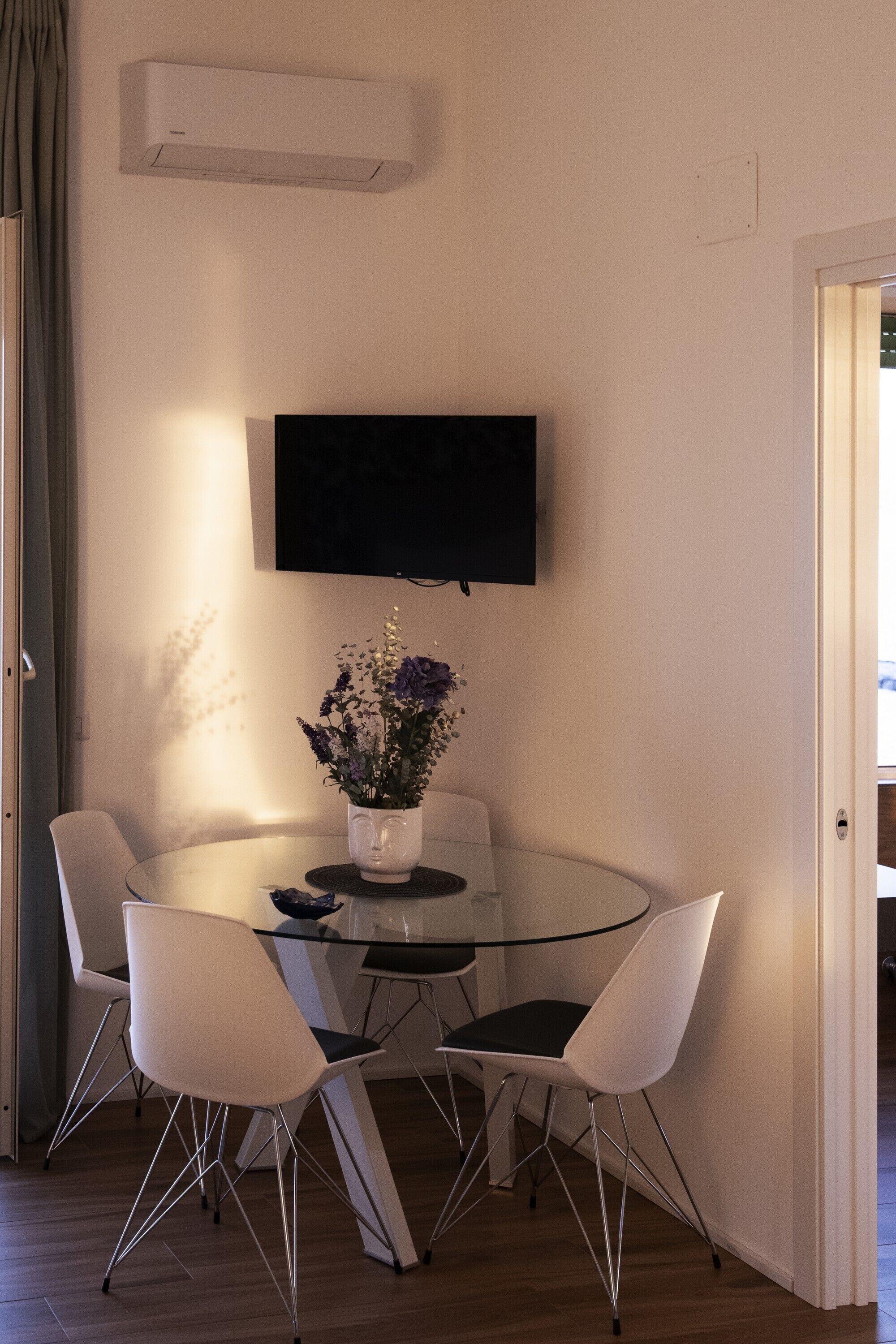 Een eetgedeelte met een glazen tafel, vier witte stoelen, een vaas met bloemen, een wandgemonteerde flatscreen-tv en een airconditioning op de muur.