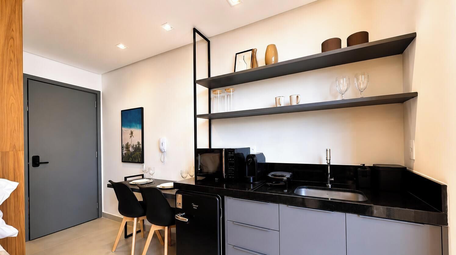 A modern kitchen with black countertops, a refrigerator, a microwave, and a sink. There are two black chairs and a dining table.