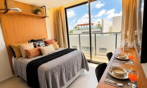 Une chambre d’hôtel avec un lit, une table à manger dressée pour deux personnes et une grande fenêtre donnant sur le paysage urbain.