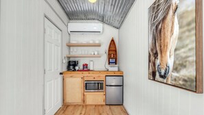 Eine kompakte Küche mit Holzausstattung, einer Mikrowelle und einem Kühlschrank. Ein Holzregal beherbergt Tassen und eine Kaffeemaschine. An der Wand hängt ein Bild eines Pferdes.